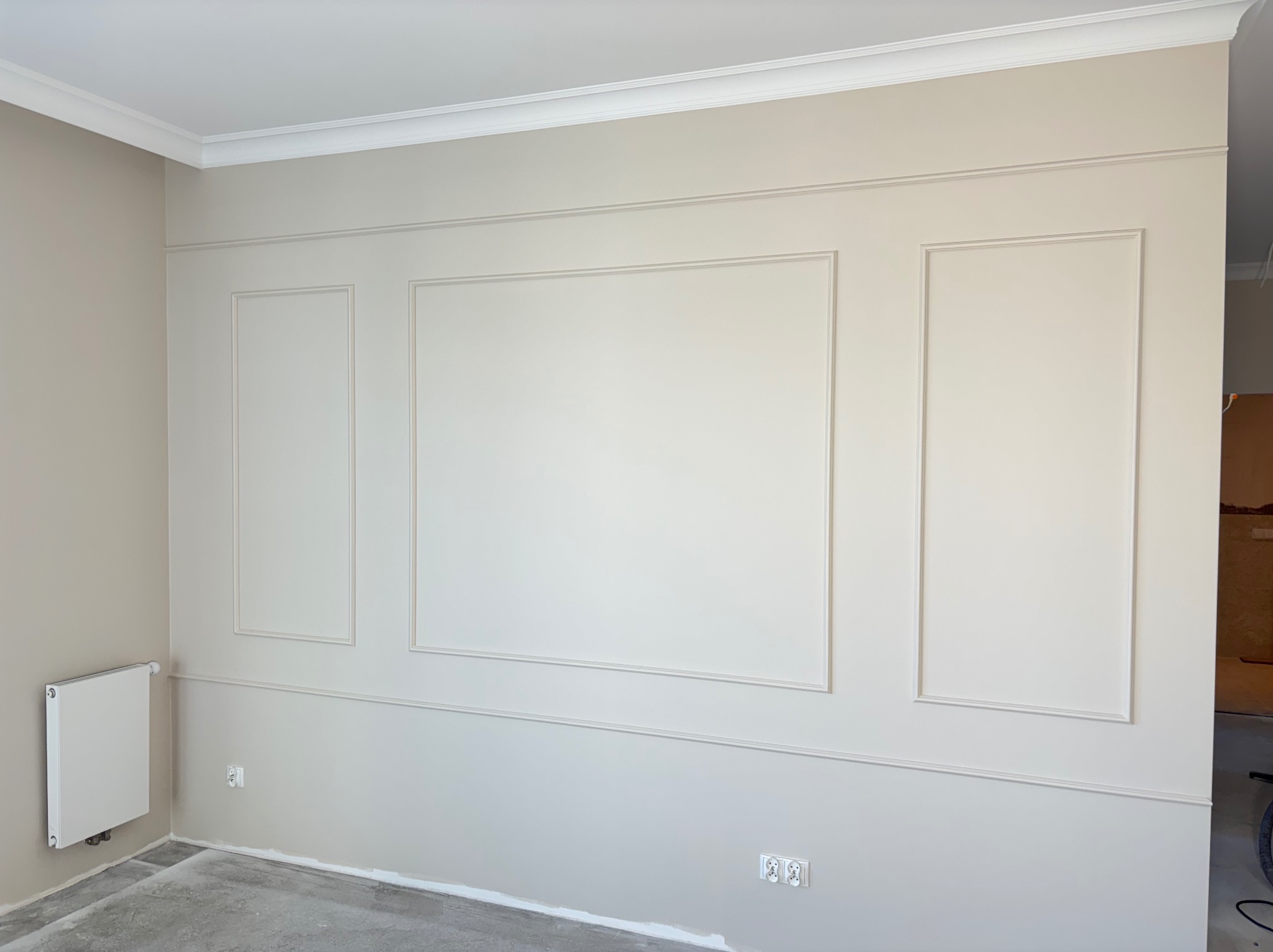 Eleganckie wykończenie ścian z dekoracyjnymi panelami i listwami w odcieniach beżu i bieli. Widoczny grzejnik i surowa podłoga przed wykończeniem.