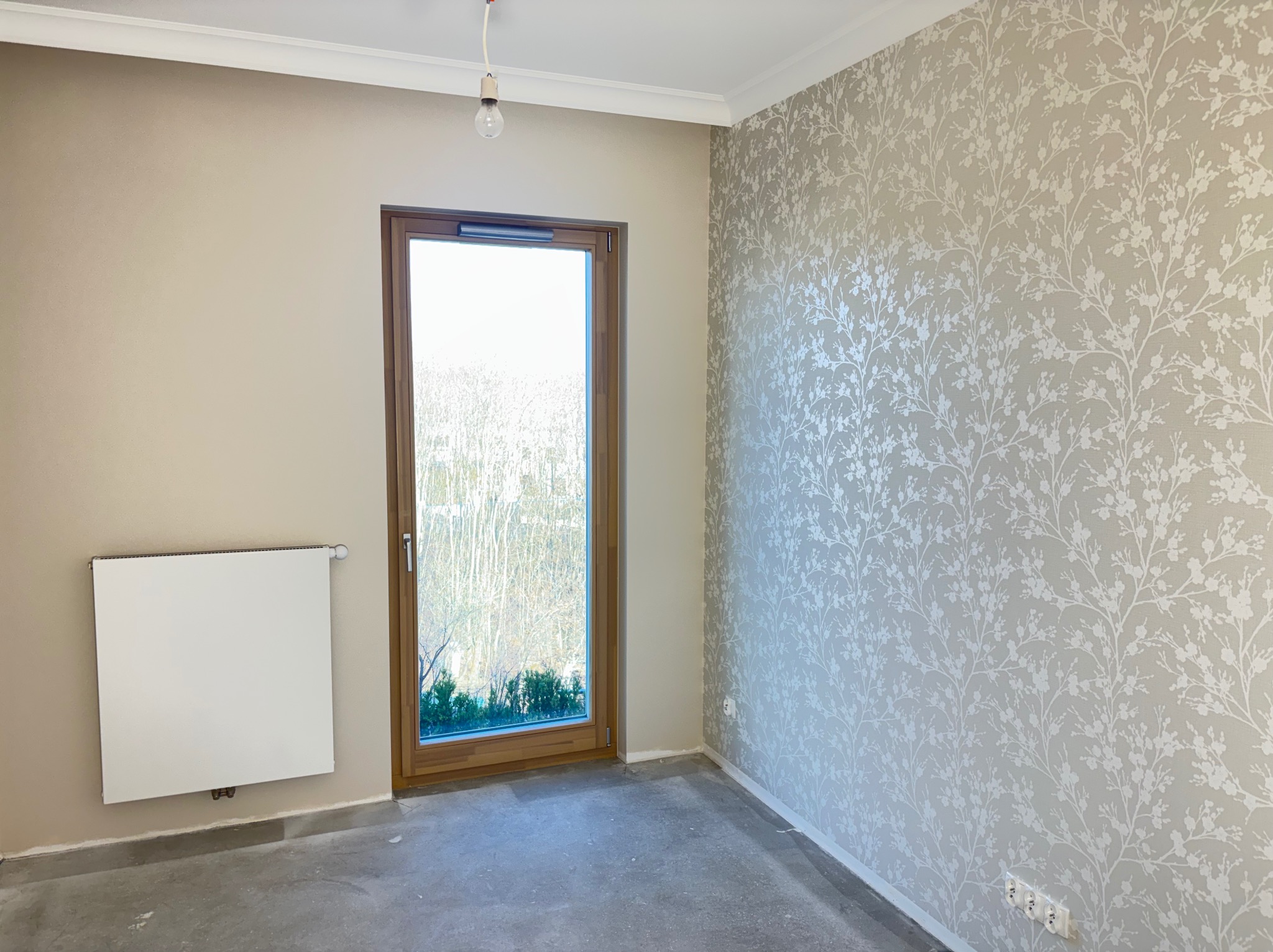 Pokój po remoncie: beżowa ściana, okno z widokiem na zieleń, dekoracyjna tapeta w kwiaty na ścianie, biały grzejnik. Surowa podłoga przed położeniem paneli.