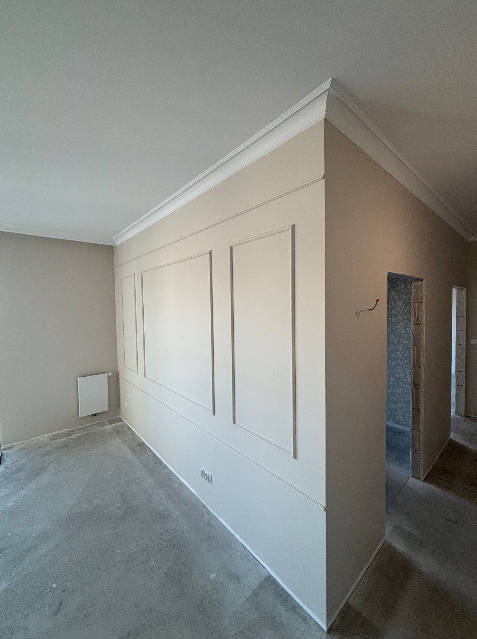 Pokój po remoncie z dekoracyjnymi panelami ściennymi w odcieniach beżu i bieli, widoczne gniazdka elektryczne i surowa podłoga. W tle widoczne kolejne pomieszczenia.