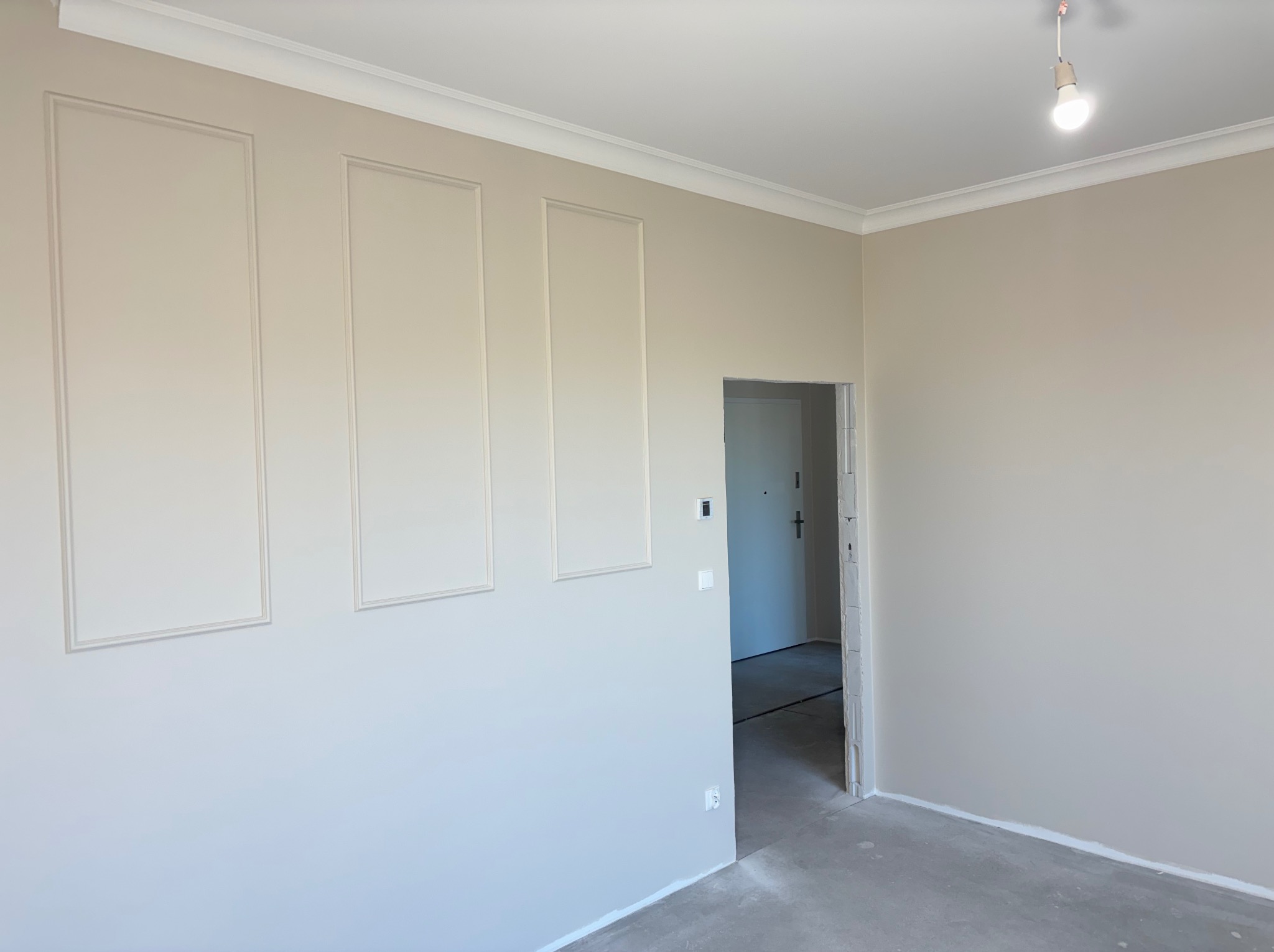 Jasne wnętrze z dekoracyjnymi panelami ściennymi i listwami przysufitowymi. Widoczny surowy beton na podłodze i otwarte przejście do innego pomieszczenia.