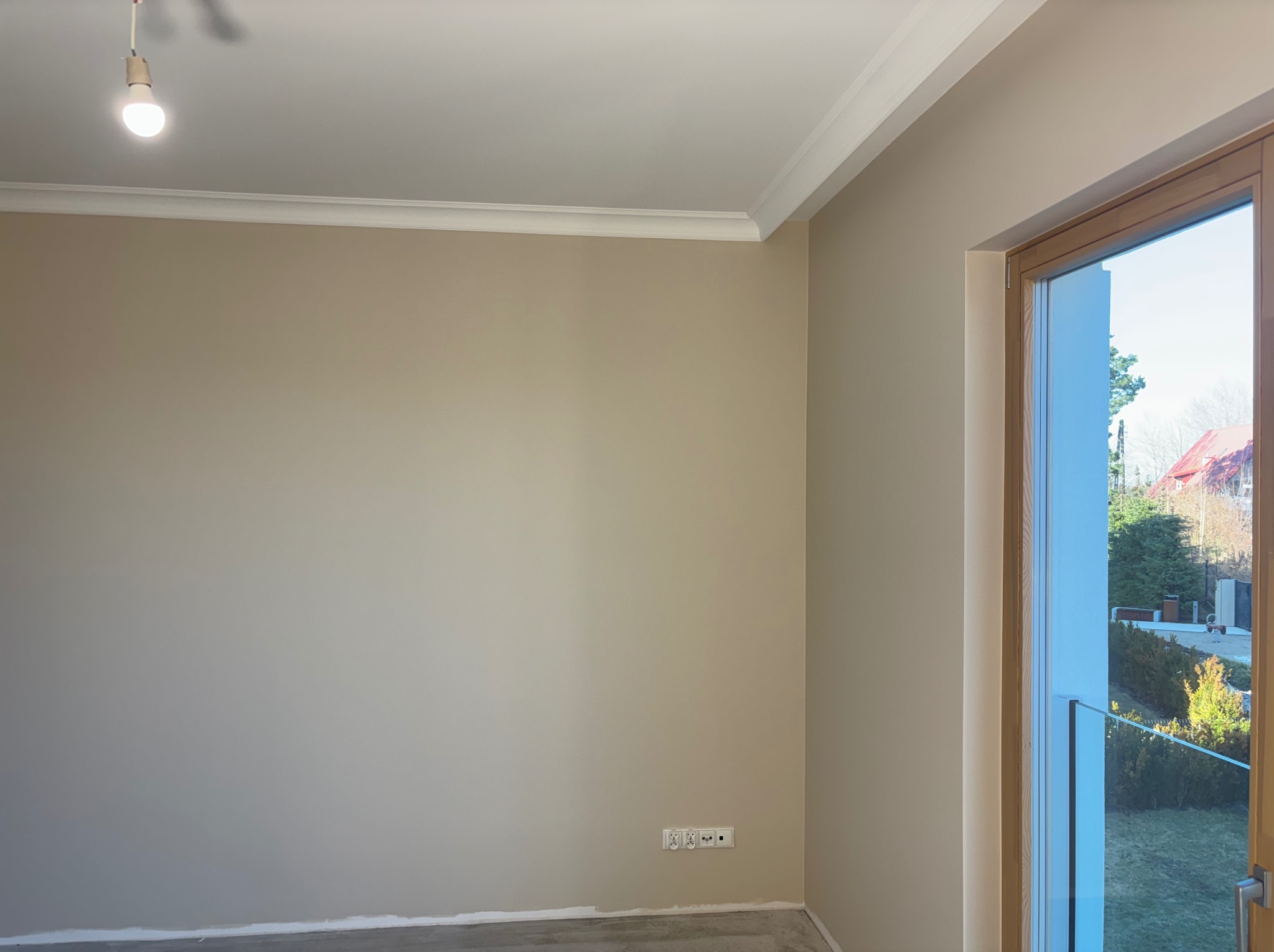 Beżowa ściana po malowaniu z białym sztukateryjnym wykończeniem sufitu i drewnianą ramą okienną. Widoczne gniazdka elektryczne i żarówka.