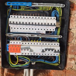USŁUGI BUDOWLANE POMIARY ELEKTRYCZNE ANDRZEJ ZIMNY - Otwarta skrzynka rozdzielcza z rzędami białych wyłączników Legrand i pomarańczowych Simtec, starannie ułożonymi niebieskimi, żółto-zielonymi i brązowymi przewodami na tle ceglanej ściany.