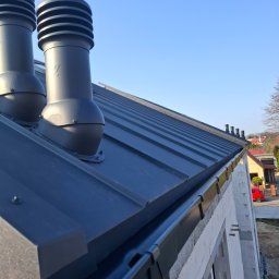 PAKODACH Piotr Toruńczak - Nowoczesny, grafitowy dach z blachy na budynku w trakcie budowy, z widocznymi kominkami wentylacyjnymi i rynnami. Ujęcie z dołu, błękitne niebo w tle.