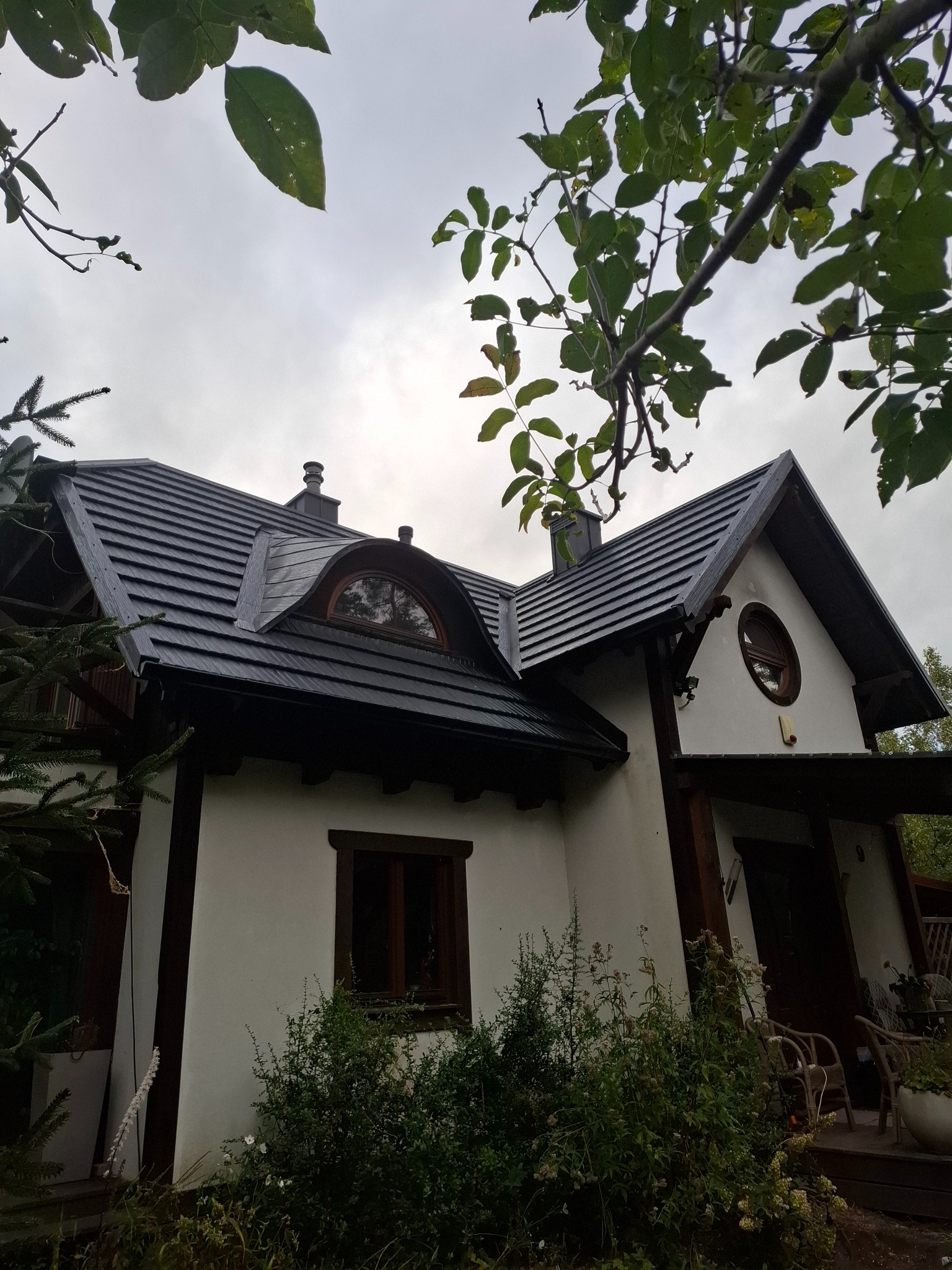 Dom z ciemną dachówką karpiówką i oknami w drewnianych ramach, widziany z dołu przez gałęzie drzew. Nietypowe okno w kształcie półkola na poddaszu.