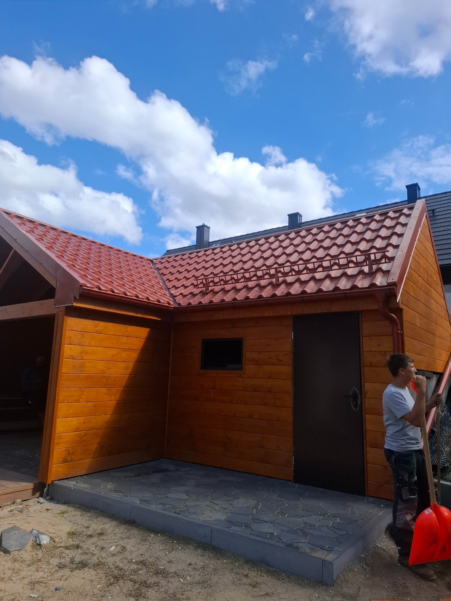 Drewniany budynek gospodarczy z nowym, czerwonym dachem z blachodachówki, rynnami i obróbkami. Na pierwszym planie mężczyzna z łopatą.