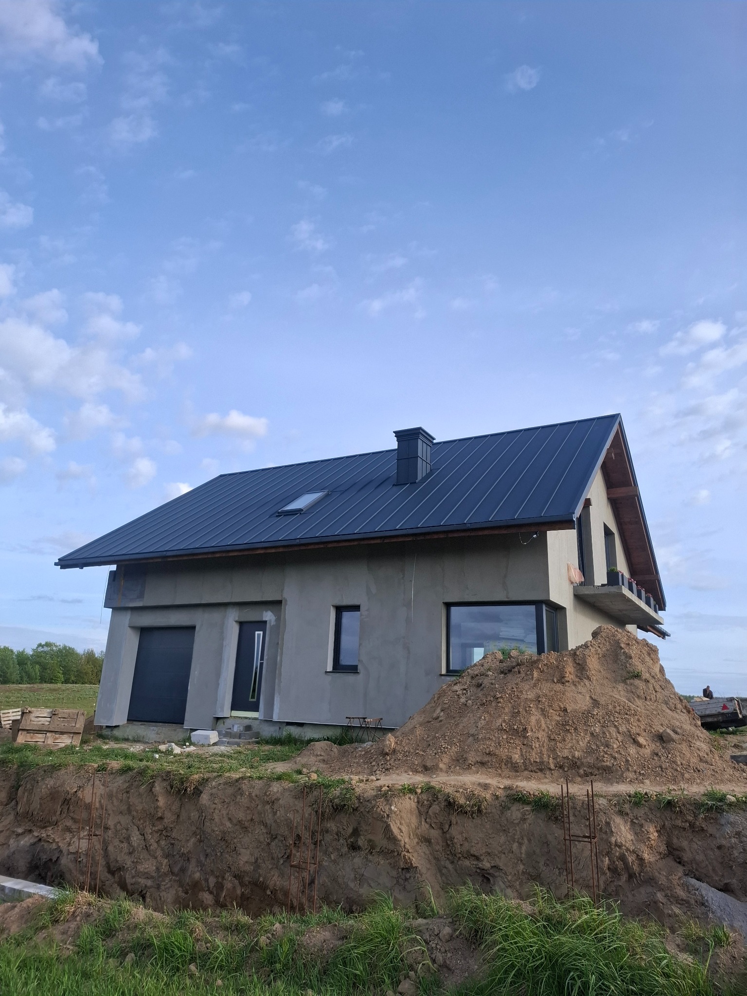 Dom w stanie surowym zamkniętym z ciemnym dachem w Załakowie. Widoczny fragment wykopu i sterta ziemi. Dach z blachy z kominem i oknem dachowym.