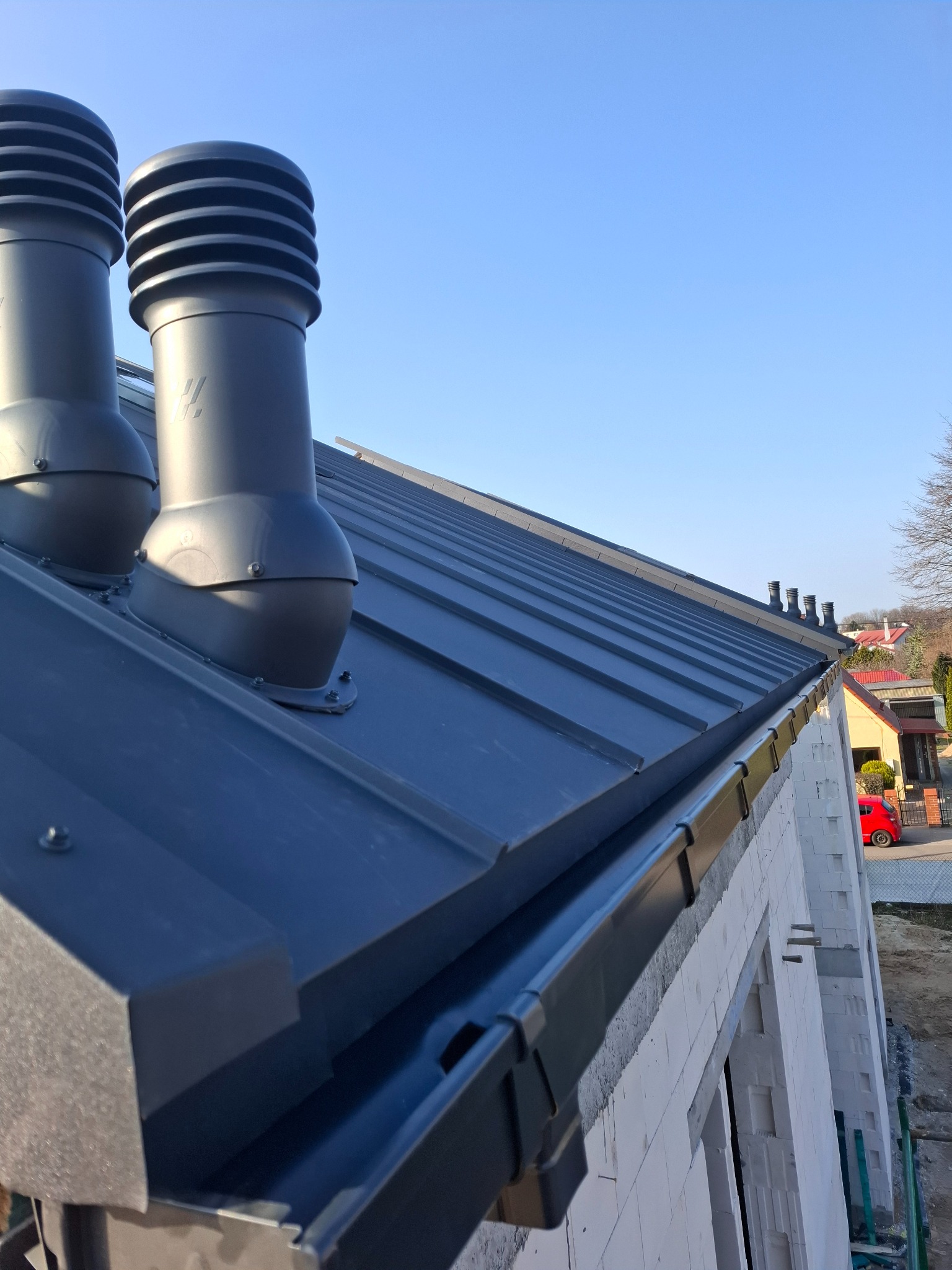 Nowoczesny, grafitowy dach z blachy na budynku w trakcie budowy, z widocznymi kominkami wentylacyjnymi i rynnami. Ujęcie z dołu, błękitne niebo w tle.