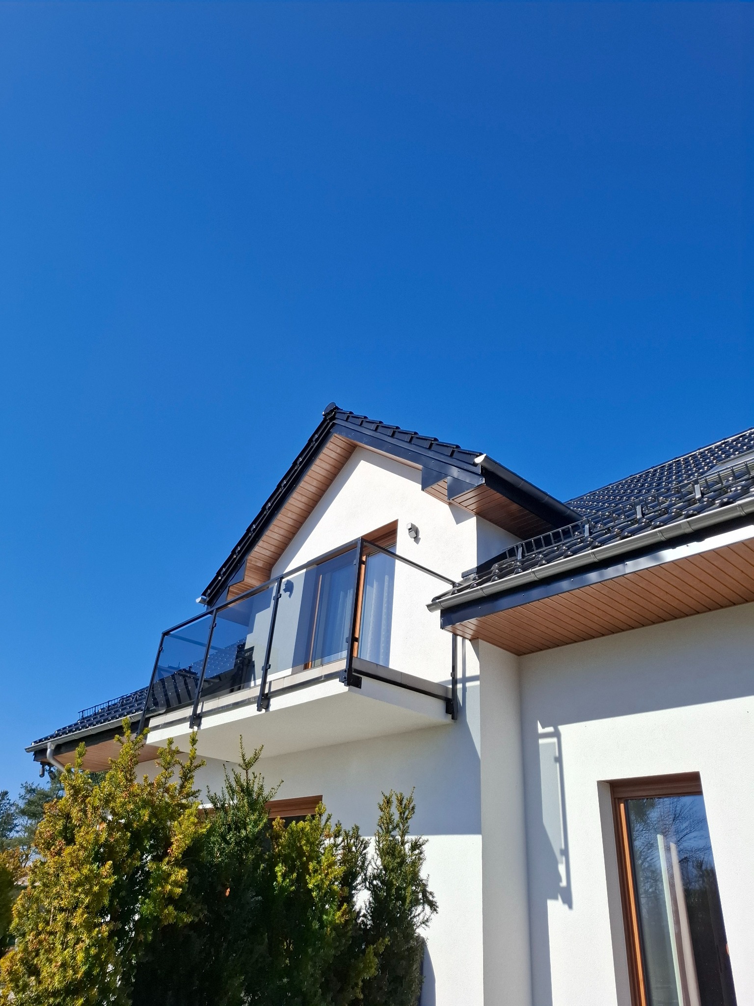 Biały dom z balkonem z przeszkloną balustradą i ciemną dachówką na tle błękitnego nieba. Widoczne okno i fragment zieleni.