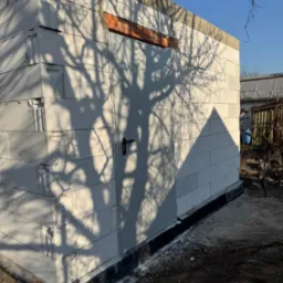 Niski budynek z białych bloczków, z widocznymi spoinami i drzwiami, na które pada cień drzewa. W tle widoczny ogród i błękitne niebo.