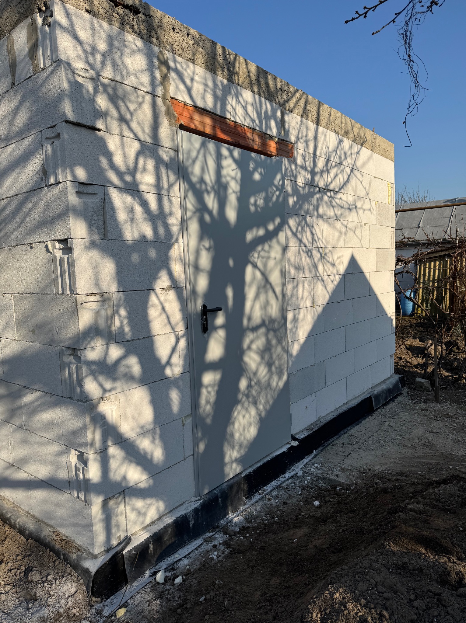 Niski budynek z białych bloczków, z widocznymi spoinami i drzwiami, na które pada cień drzewa. W tle widoczny ogród i błękitne niebo.