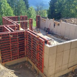 Konstrukt - Fundamenty domu jednorodzinnego z widocznym szalunkiem systemowym PERI, częściowo wylane betonem, otoczone zielenią i ziemią.