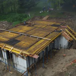 Wysoki kąt widzenia na budowę domu. Widoczne szalunki i zbrojenie przygotowane pod wylewkę stropu. W tle zalesiony teren i pracownicy budowlani.