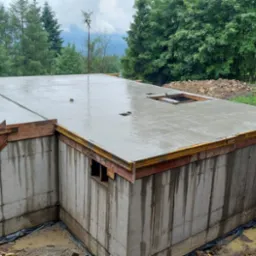 Świeżo wylany, mokry beton na stropie i schodach piwnicy budynku w surowym stanie, otoczony drewnianym szalunkiem, widoczne ślady wody na betonowych ścianach piwnicy, wykop ziemny wokół fundamentów.