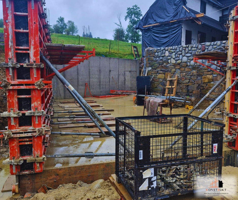 Budowa fundamentów w terenie podmokłym, widoczne szalunki i stalowe podpory, w tle dom z kamienną elewacją częściowo przykryty folią.