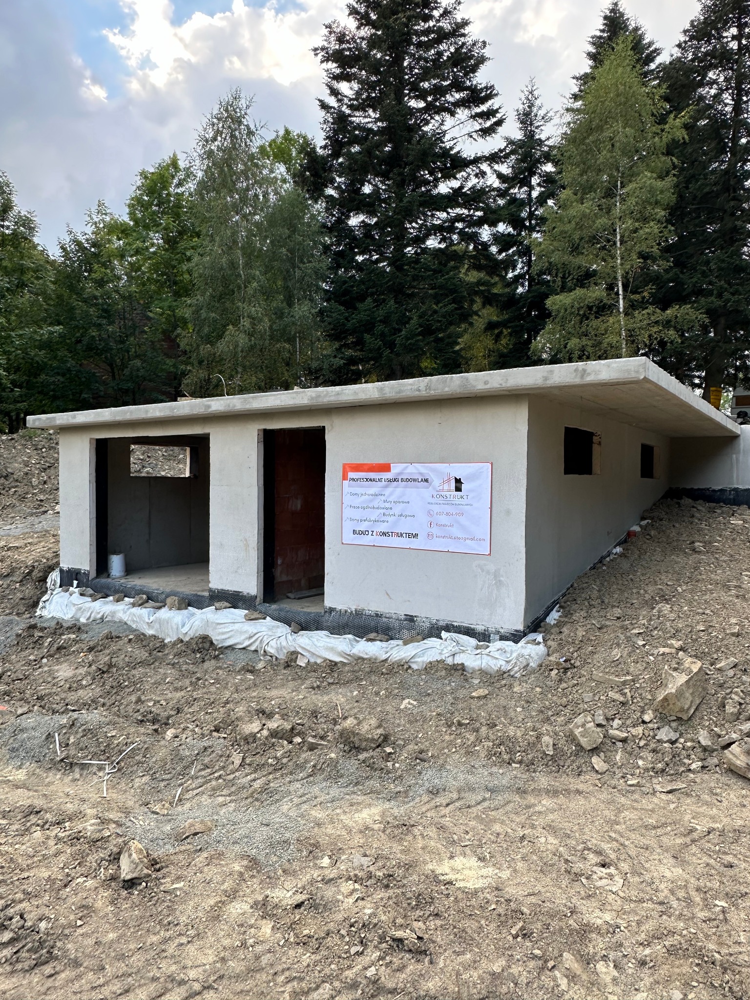 Surowy budynek z betonu w trakcie budowy, z widoczną konstrukcją fundamentów i ścian, otoczony ziemią i drzewami, z plakatem reklamowym firmy budowlanej na ścianie.