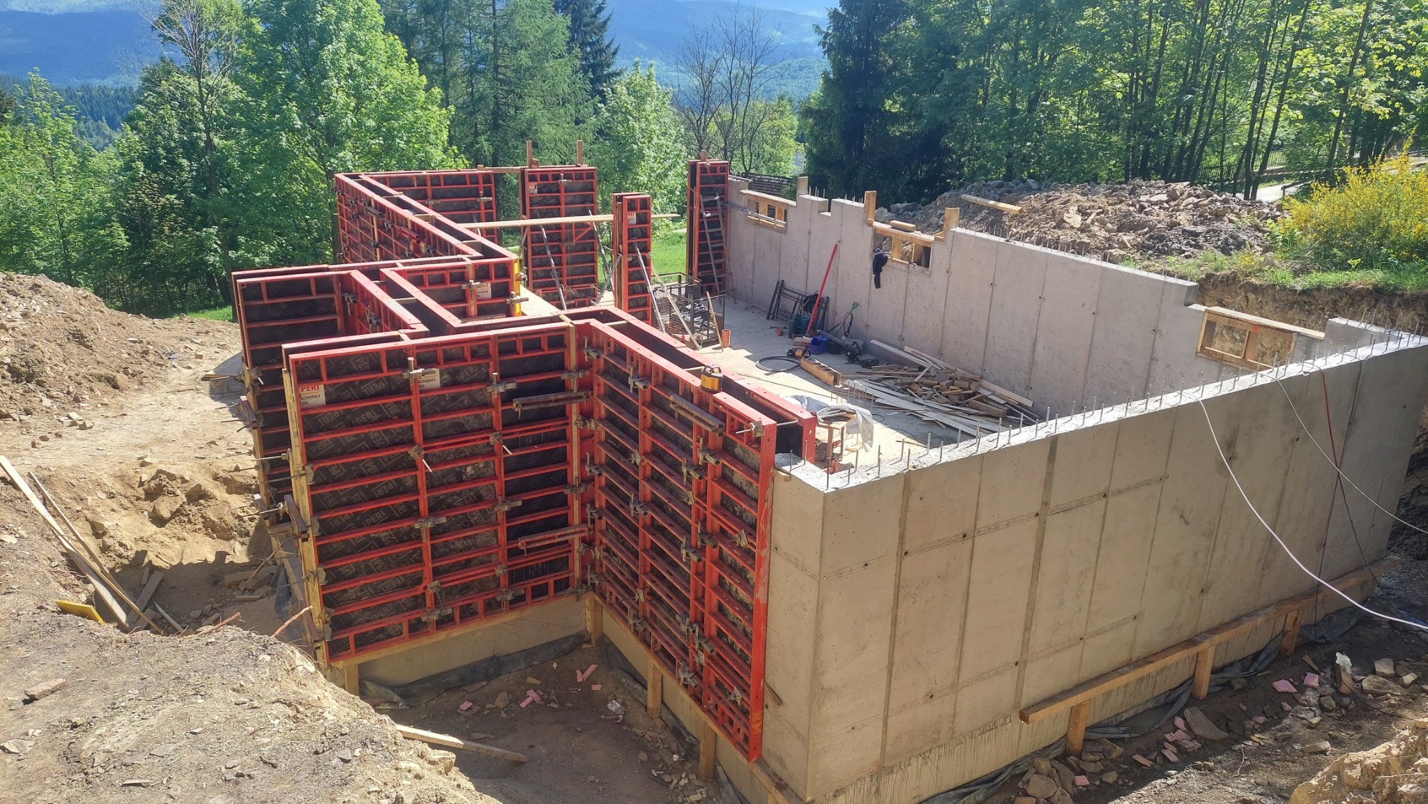 Fundamenty domu jednorodzinnego z widocznym szalunkiem systemowym PERI, częściowo wylane betonem, otoczone zielenią i ziemią.