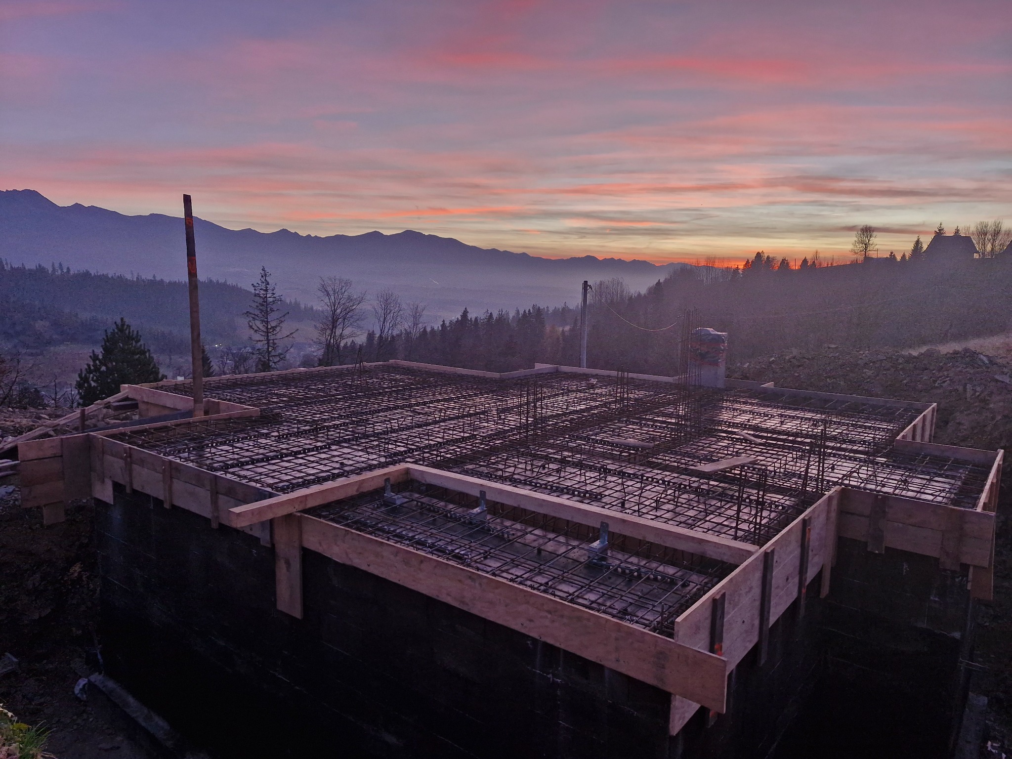 Siatka zbrojeniowa ułożona w szalunku fundamentu na tle malowniczego, wschodzącego słońca nad górami.