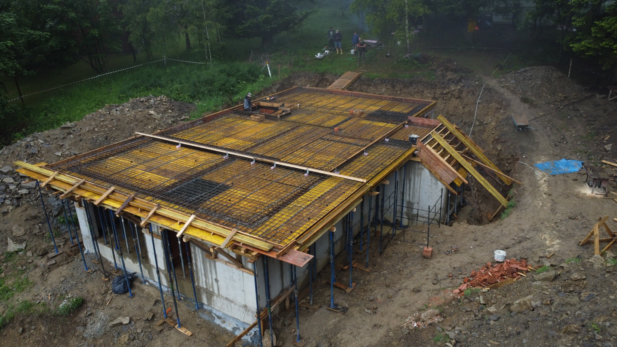 Wysoki kąt widzenia na budowę domu. Widoczne szalunki i zbrojenie przygotowane pod wylewkę stropu. W tle zalesiony teren i pracownicy budowlani.