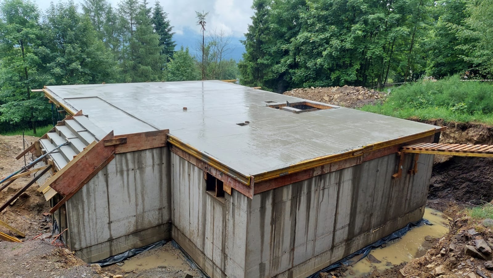 Świeżo wylany, mokry beton na stropie i schodach piwnicy budynku w surowym stanie, otoczony drewnianym szalunkiem, widoczne ślady wody na betonowych ścianach piwnicy, wykop ziemny wokół fundamentów.