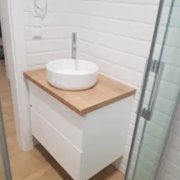 Nowoczesna łazienka z białą umywalką nablatową na drewnianym blacie i białymi płytkami typu metro na ścianach, widoczna kabina prysznicowa z chromowanymi okuciami.