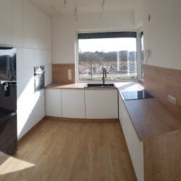 WOODLUCK - Nowoczesna, jasna kuchnia z białymi szafkami i drewnianym blatem, czarny zlew pod oknem z widokiem na krajobraz za oknem, czarna lodówka po lewej stronie, brak wykończenia sufitu.