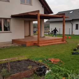 WOODLUCK - Drewniana altana z tarasem w trakcie budowy przy domu jednorodzinnym, widoczny stolarz przy pracy i narzędzia, ogródek warzywny w pierwszym planie, pochmurne niebo.