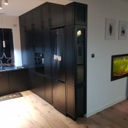 WOODLUCK - Czarne meble kuchenne na wymiar z lodówką w zabudowie i szklanymi witrynami, oświetlenie punktowe, drewniana podłoga.