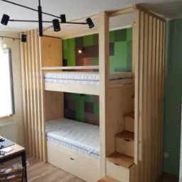 Drewniane łóżko piętrowe z wbudowanymi schodkami i szufladami, z pionowymi listwami dekoracyjnymi, na tle ściany z motywem Minecrafta, oświetlone nowoczesną lampą sufitową.