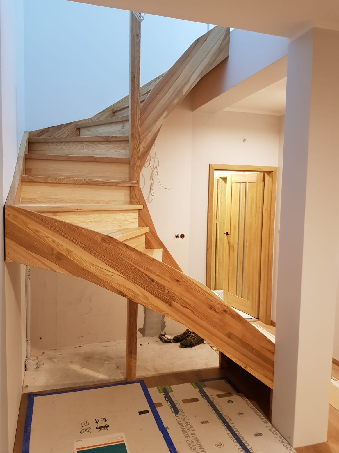 Jasne, drewniane schody z poręczą, częściowo ukończone, z widocznymi elementami konstrukcyjnymi i zabezpieczeniami podłogi, prowadzące do drewnianych drzwi w tle.