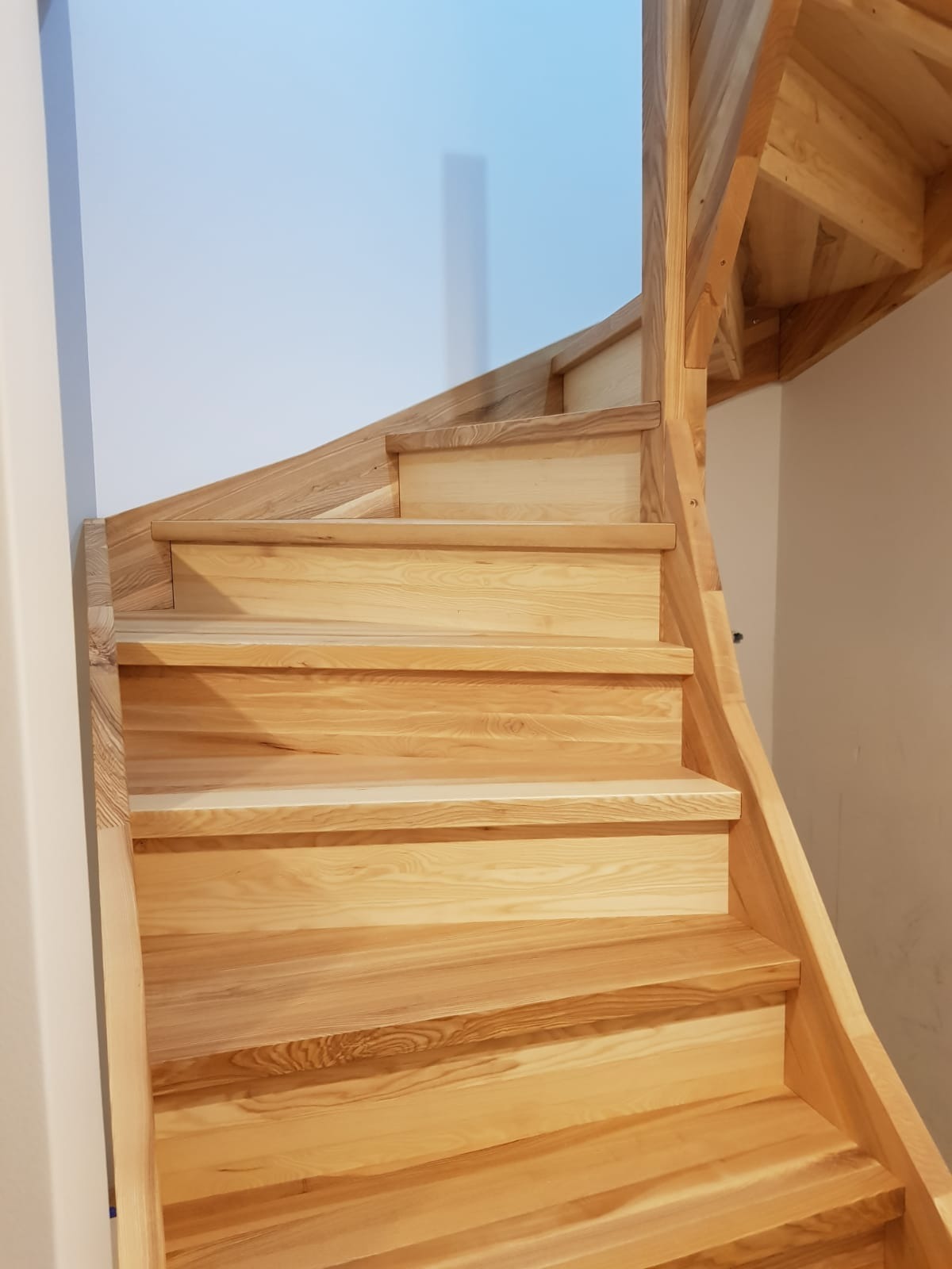 Spiralne schody drewniane z jasnego drewna, widok z dołu, naturalne usłojenie drewna, białe ściany w tle.