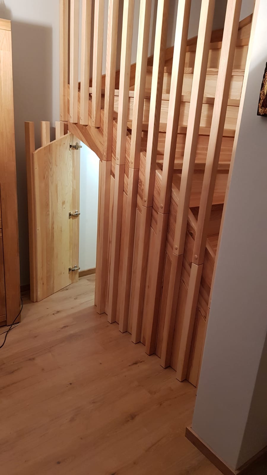 Schody drewniane z pionowymi szczeblami i ukrytym wejściem pod nimi, zabezpieczonym drzwiczkami z zawiasami.