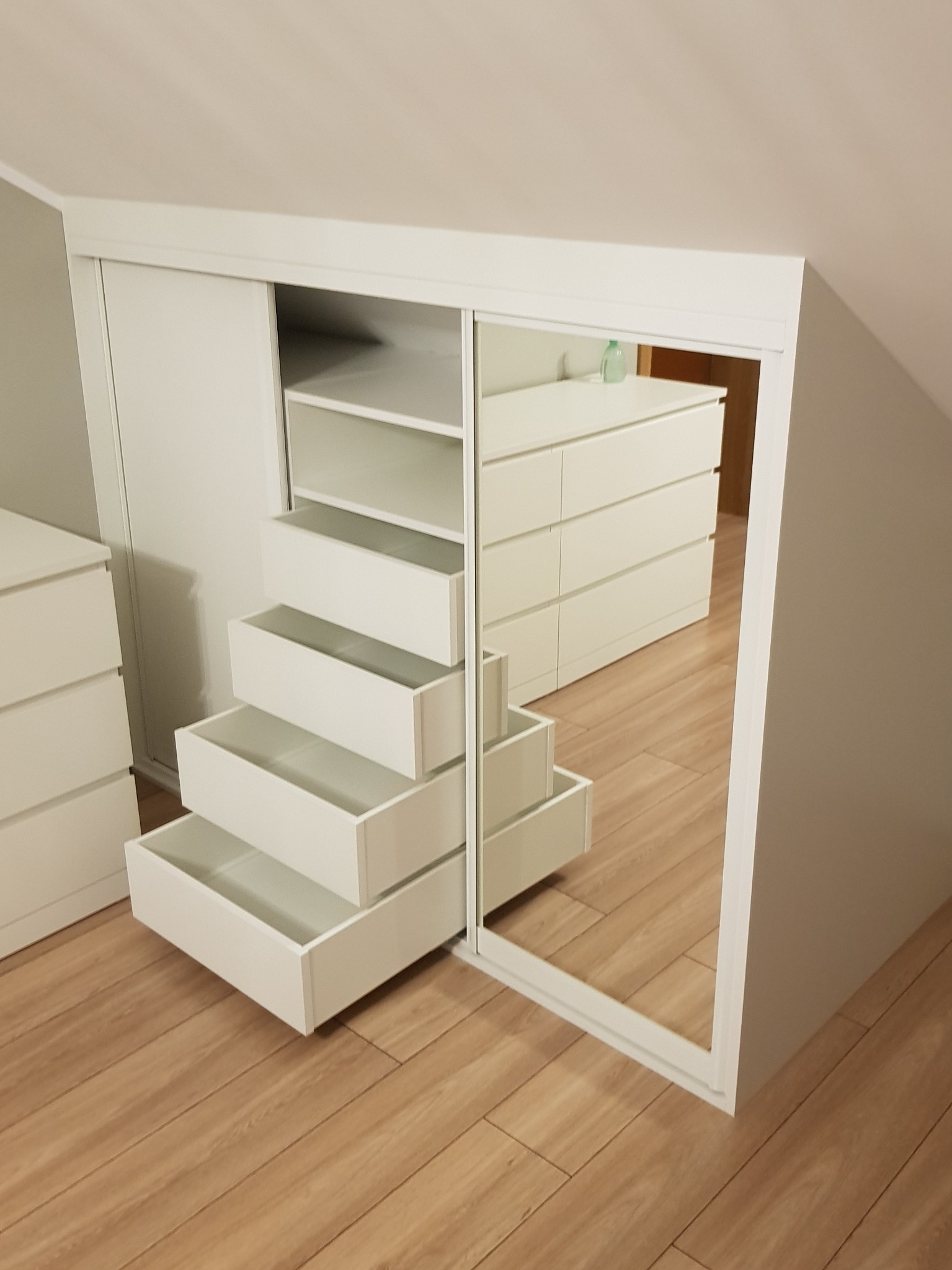 Biała szafa wnękowa z lustrzanymi drzwiami przesuwnymi i otwartymi szufladami w kształcie schodków, widoczna na tle poddasza z drewnianą podłogą i komodą.