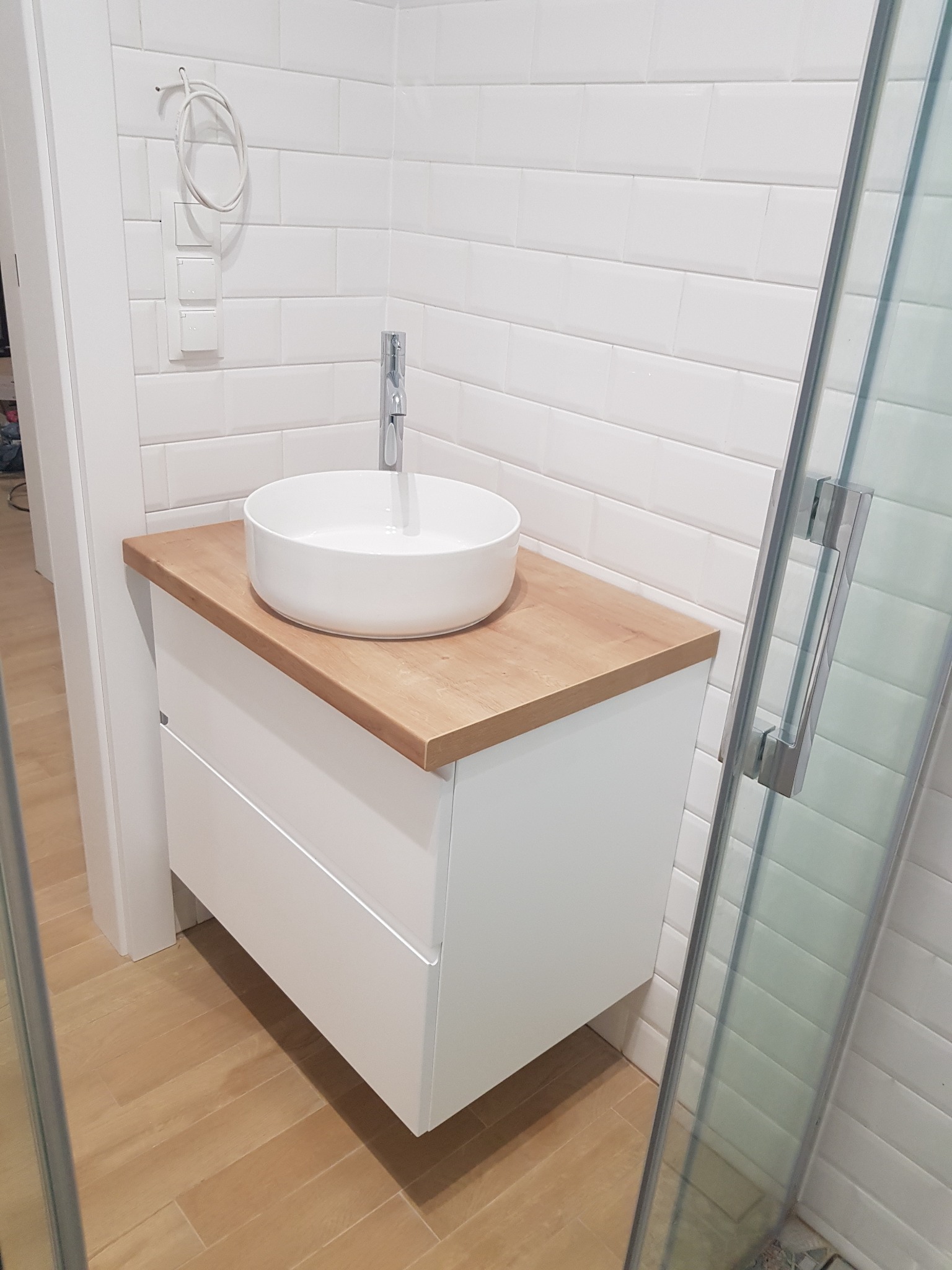 Nowoczesna łazienka z białą umywalką nablatową na drewnianym blacie i białymi płytkami typu metro na ścianach, widoczna kabina prysznicowa z chromowanymi okuciami.