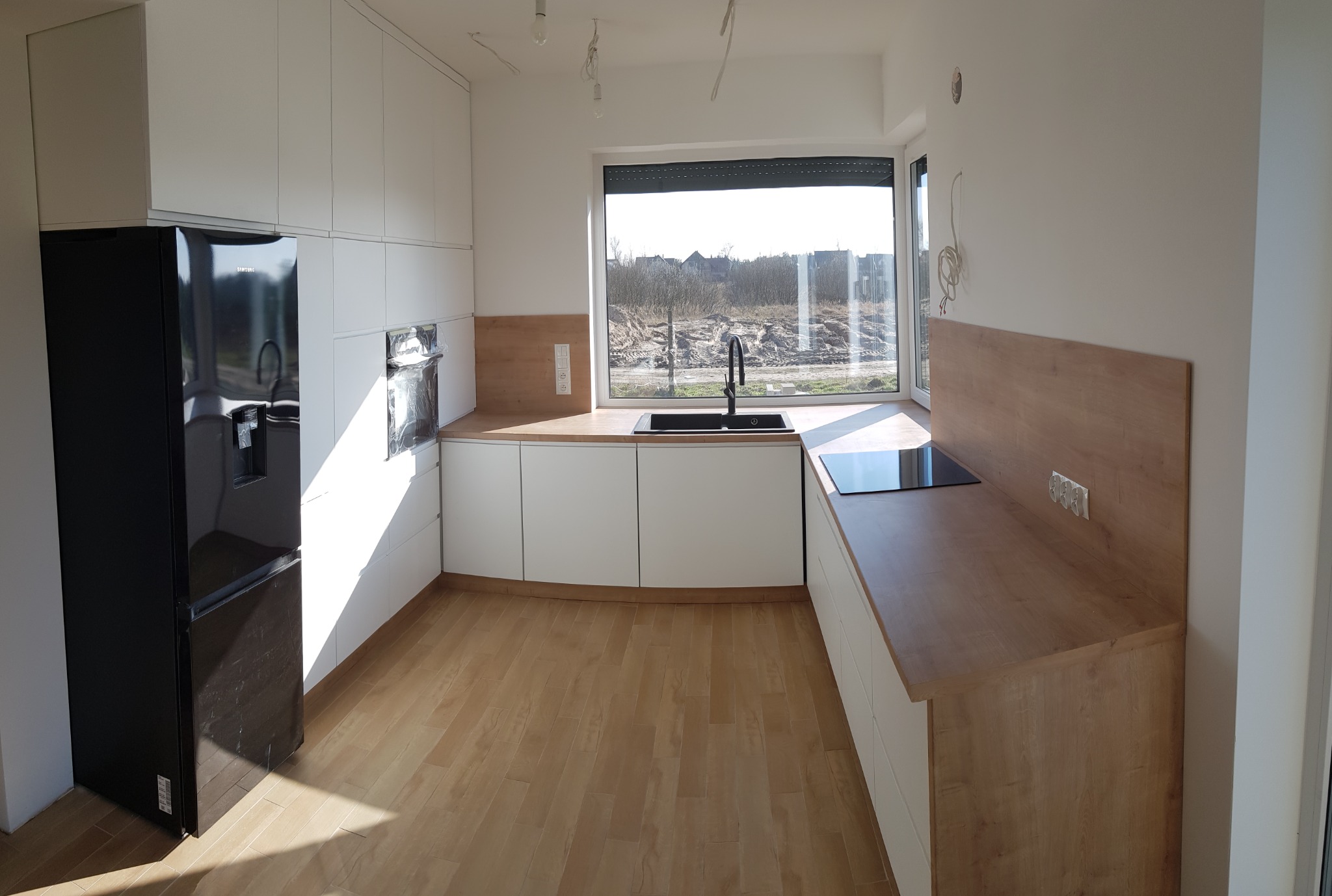 Nowoczesna, jasna kuchnia z białymi szafkami i drewnianym blatem, czarny zlew pod oknem z widokiem na krajobraz za oknem, czarna lodówka po lewej stronie, brak wykończenia sufitu.