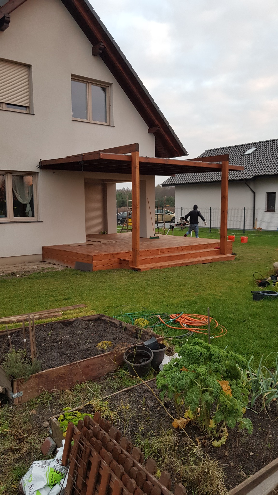 Drewniana altana z tarasem w trakcie budowy przy domu jednorodzinnym, widoczny stolarz przy pracy i narzędzia, ogródek warzywny w pierwszym planie, pochmurne niebo.