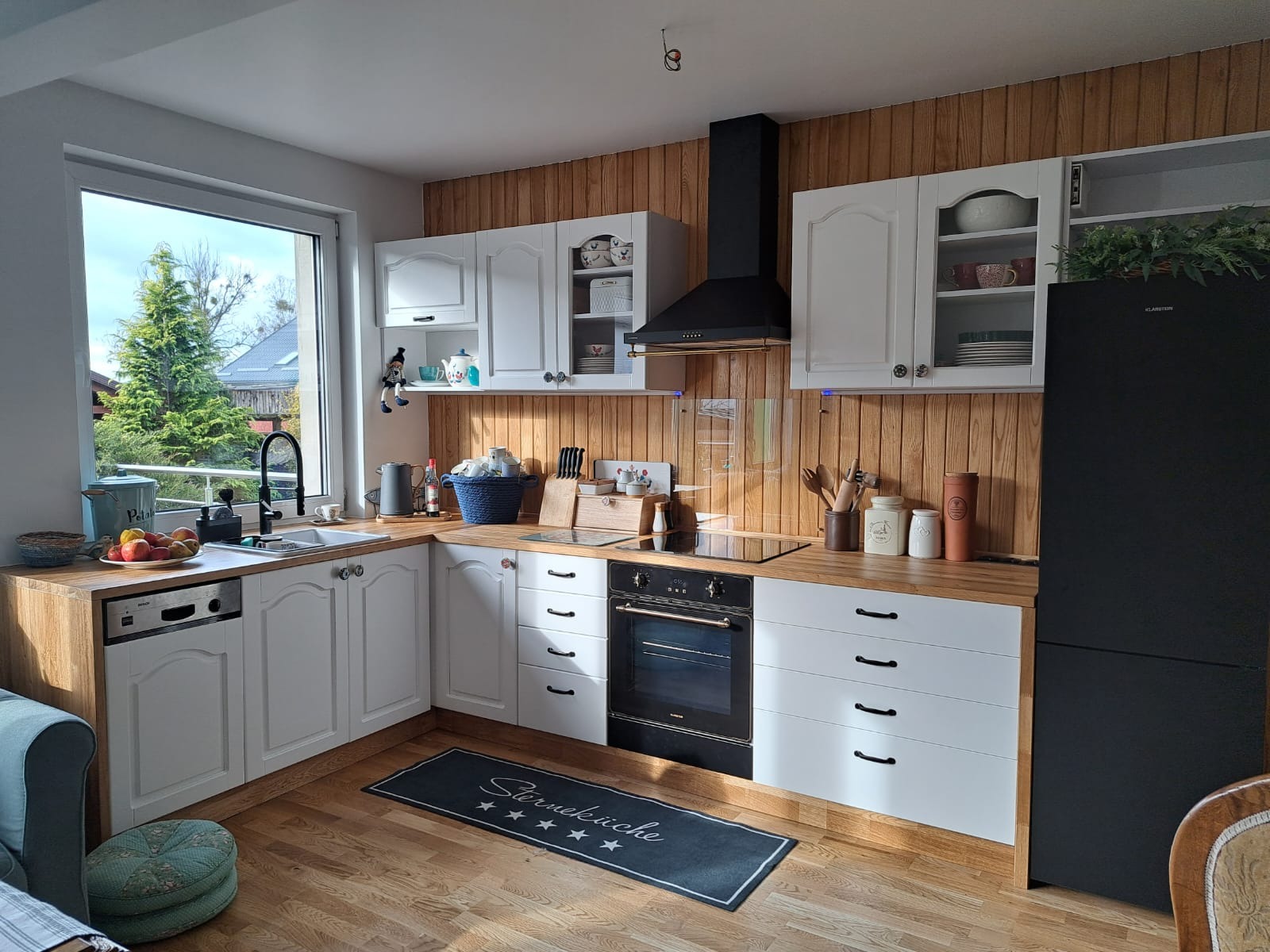 Jasna kuchnia z drewnianymi akcentami, białe szafki z frezowanymi frontami, drewniane blaty, czarny piekarnik i lodówka, widok na ogród przez okno, mata podłogowa z napisem.
