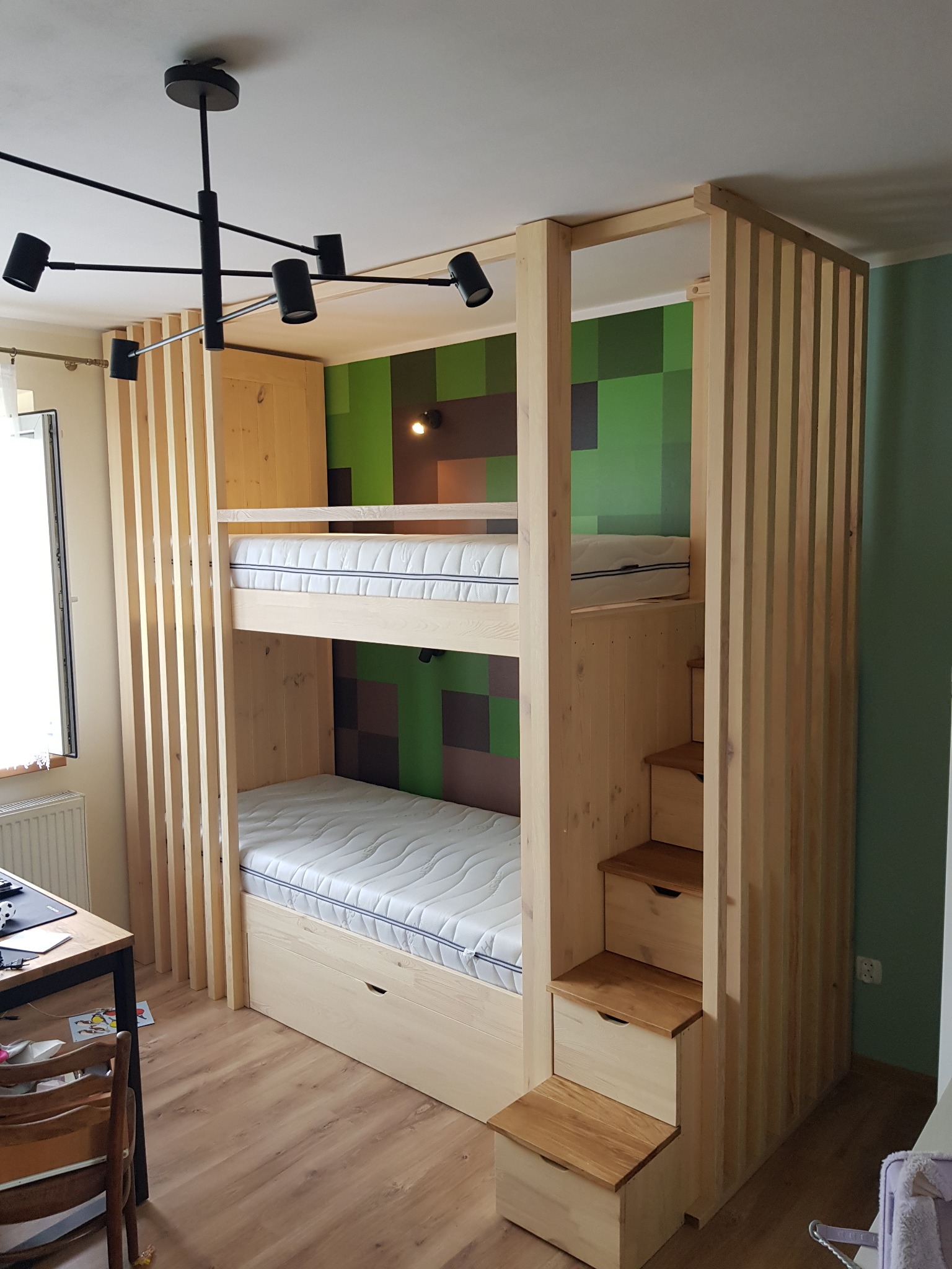 Drewniane łóżko piętrowe z wbudowanymi schodkami i szufladami, z pionowymi listwami dekoracyjnymi, na tle ściany z motywem Minecrafta, oświetlone nowoczesną lampą sufitową.