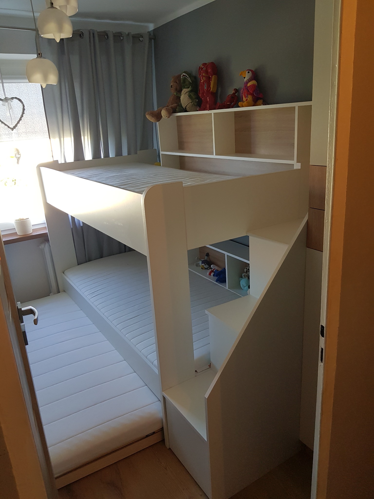 Białe łóżko piętrowe z wbudowanymi półkami i schodami, zabawki na półkach, widok z poziomu podłogi.