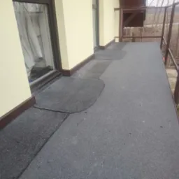 Balkon z szarą papą bitumiczną, widoczne fragmenty napraw z ciemniejszego materiału, żółta elewacja budynku z brązowymi listwami przypodłogowymi, brązowa balustrada.