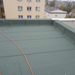 Dach-Ciekł - Zielony dach pokryty papą termozgrzewalną, widoczny palnik dekarski z pomarańczowym przewodem i pracownik w oddali, budynki w tle.