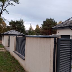 Dach-Ciekł - Nowoczesne ogrodzenie posesji z metalowymi przęsłami w kolorze grafitowym i jasnymi, otynkowanymi słupkami, widok na dom z grafitowym dachem i rynnami.