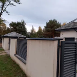 Nowoczesne ogrodzenie posesji z metalowymi przęsłami w kolorze grafitowym i jasnymi, otynkowanymi słupkami, widok na dom z grafitowym dachem i rynnami.