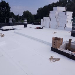 Dach-Ciekł - Białe płyty styropianowe ułożone na dachu, widoczny pracownik w trakcie pracy, narzędzia i materiały budowlane rozłożone na powierzchni dachu, w tle drzewa i niebo.