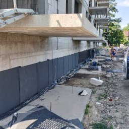 Dach-Ciekł - Czarna izolacja pionowa fundamentów budynku wielorodzinnego w trakcie realizacji, widoczna osoba i betoniarka Knauf w tle.
