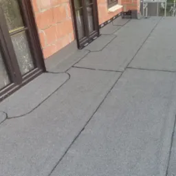 Szczegółowe ujęcie hydroizolacji na płaskim dachu z oknami, widoczne łączenia papy i elementy mocujące, ceglana ściana budynku w tle.