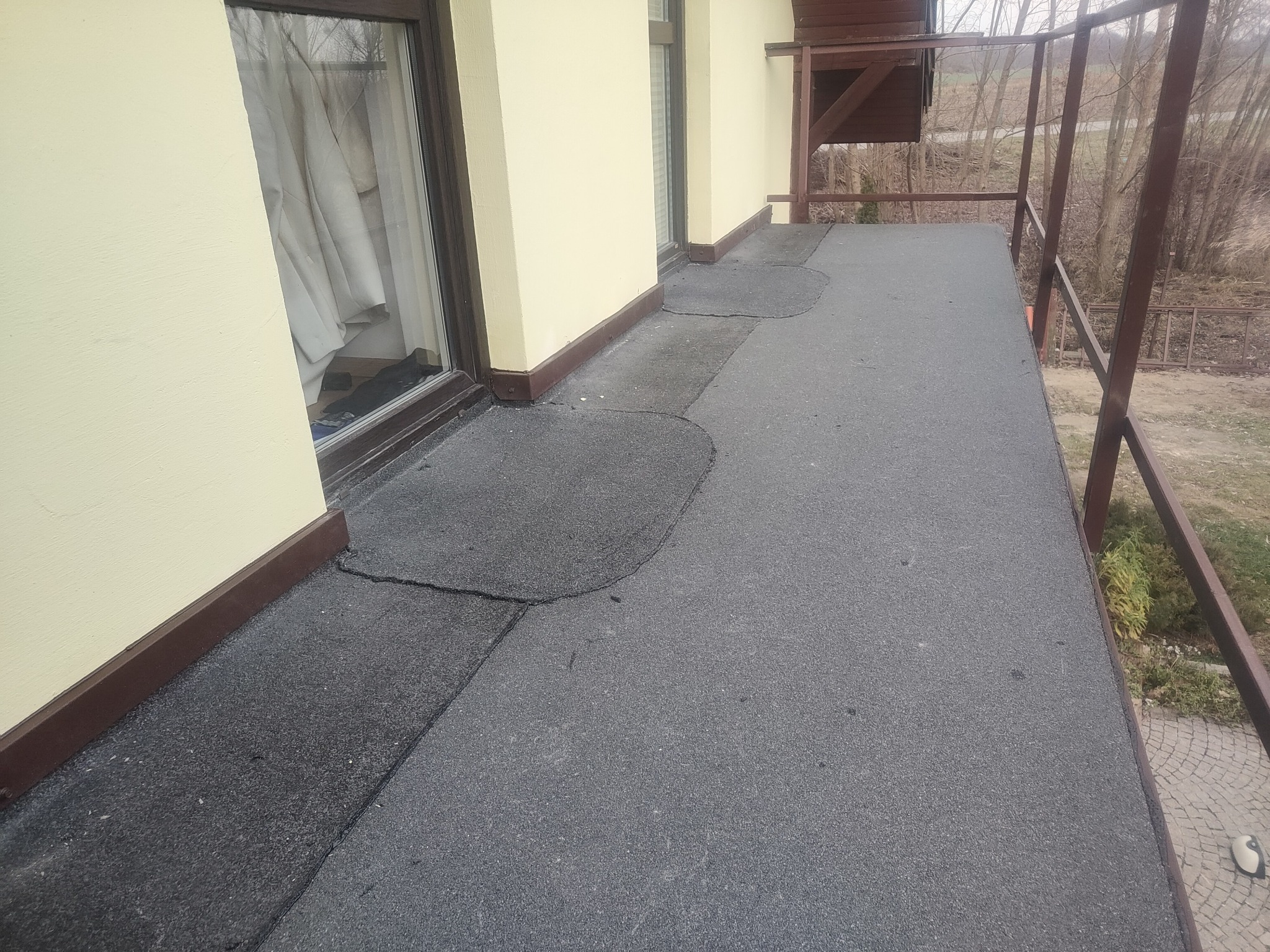 Balkon z szarą papą bitumiczną, widoczne fragmenty napraw z ciemniejszego materiału, żółta elewacja budynku z brązowymi listwami przypodłogowymi, brązowa balustrada.
