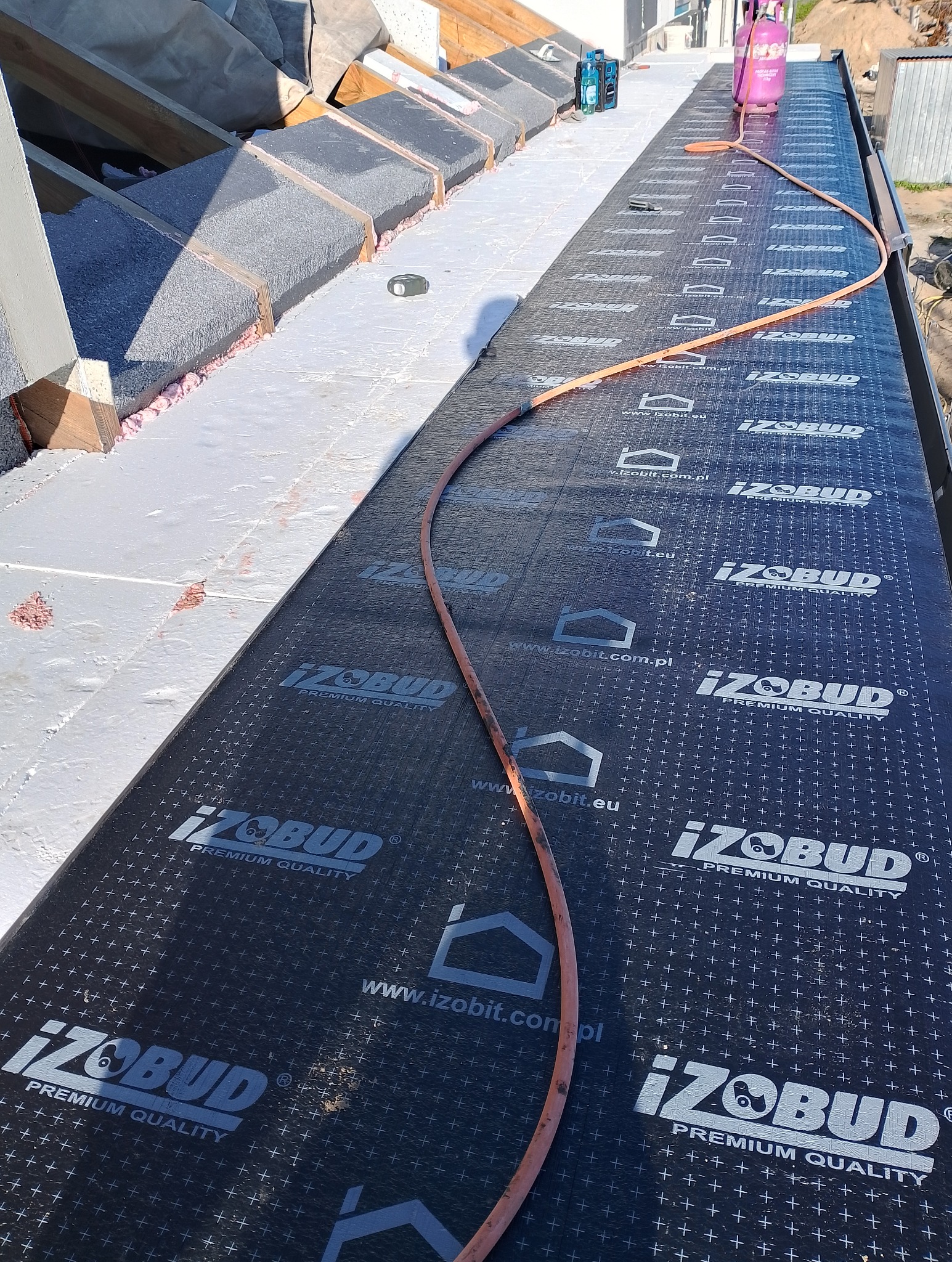 Montaż papy termozgrzewalnej IZOBUD na izolacji dachu, widoczny palnik gazowy z podłączonym wężem i butlą, w tle konstrukcja dachu z izolacją.