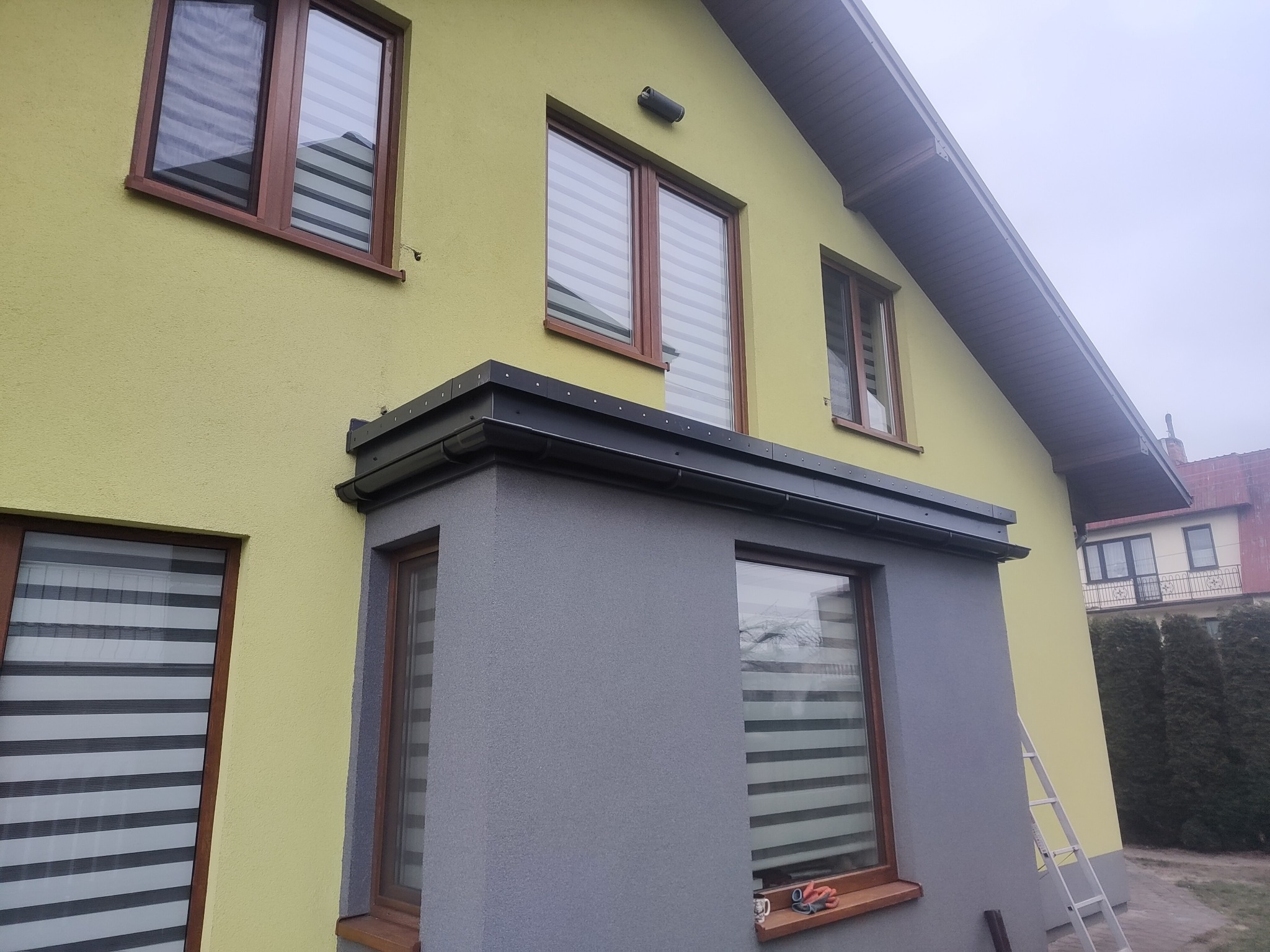 Elewacja domu z oknami z brązowymi ramami i roletami dzień-noc, fragment dachu z czarnymi obróbkami blacharskimi, ściana w dwóch odcieniach: żółtym i szarym, drabina oparta o ścianę.