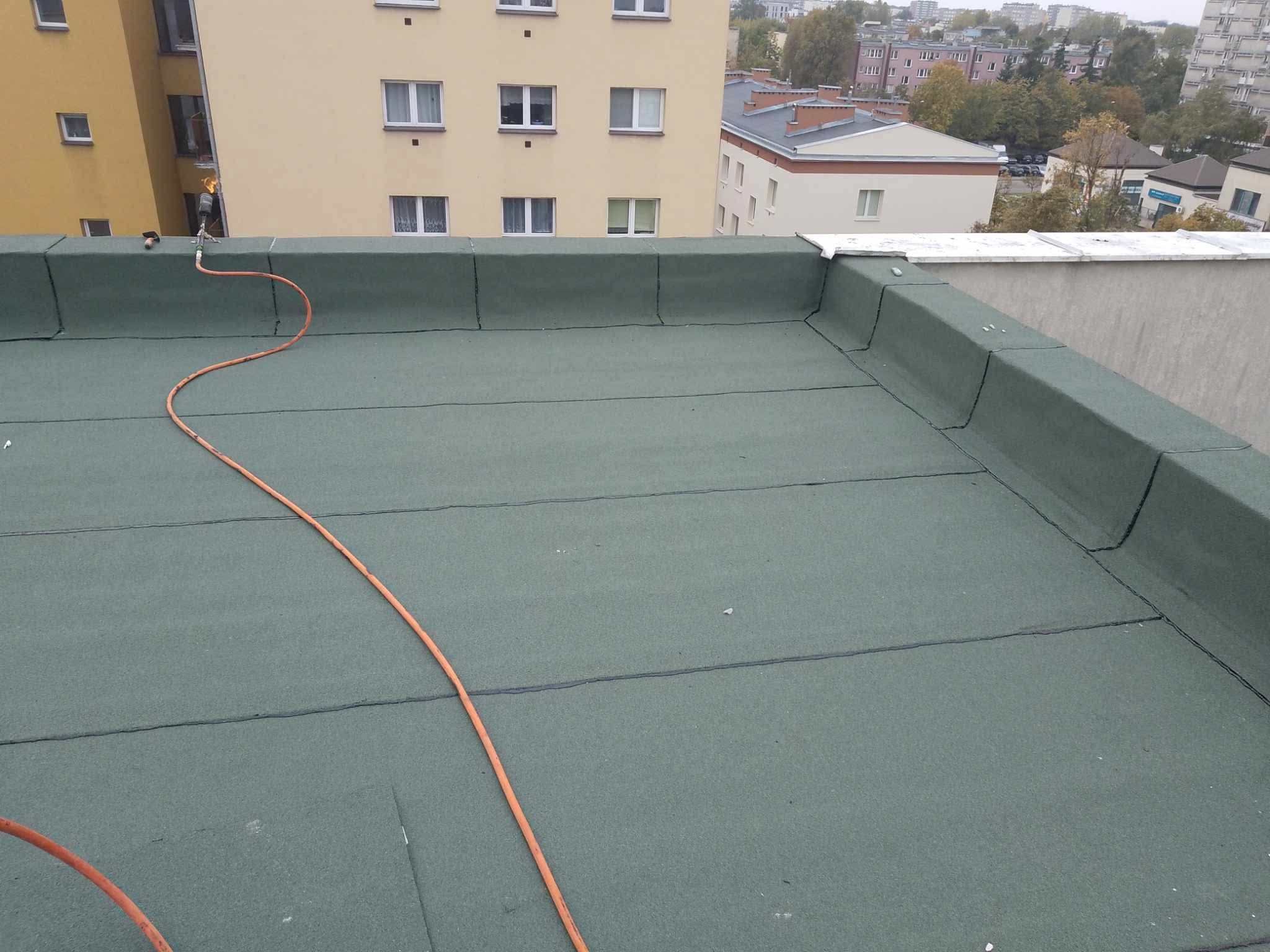 Zielony dach pokryty papą termozgrzewalną, widoczny palnik dekarski z pomarańczowym przewodem i pracownik w oddali, budynki w tle.