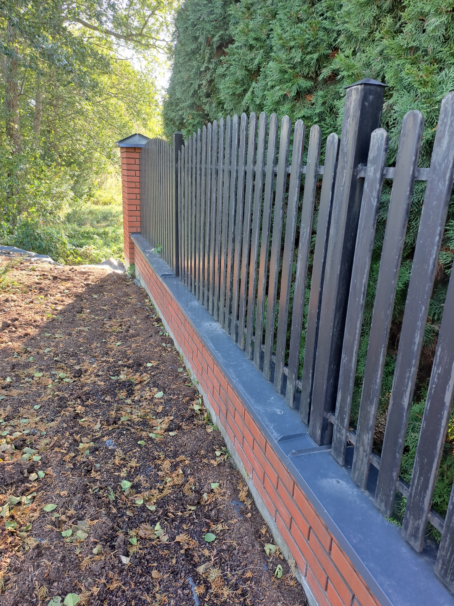 Nowo postawione ogrodzenie z ceglanymi słupkami i metalowymi przęsłami, widok z boku na tle zieleni.